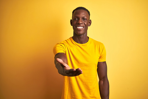 Young african american man wearing casual t-shirt standing over isolated yellow background smiling cheerful offering palm hand giving assistance and acceptance.