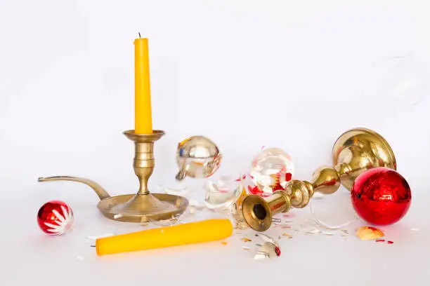 Front view of falling and shattered red and silver Christmas baubles and two bronze candleholders with a yellow candle on white background