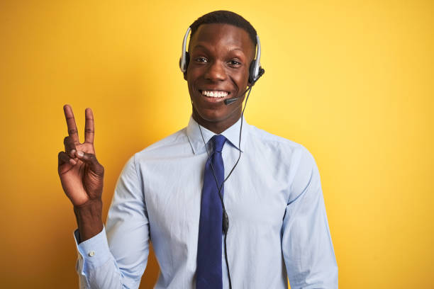 istock African american operator man working using headset over isolated yellow background smiling with happy face winking at the camera doing victory sign. Number two. 1173776995