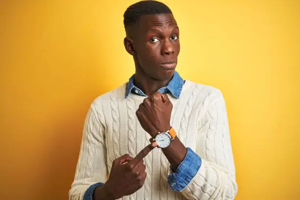 Photo of African american man wearing denim shirt and white sweater over isolated yellow background In hurry pointing to watch time, impatience, looking at the camera with relaxed expression