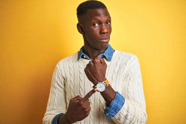 african american man wearing denim shirt and white sweater over isolated yellow background in hurry pointing to watch time, impatience, looking at the camera with relaxed expression - clock face clock deadline human hand imagens e fotografias de stock