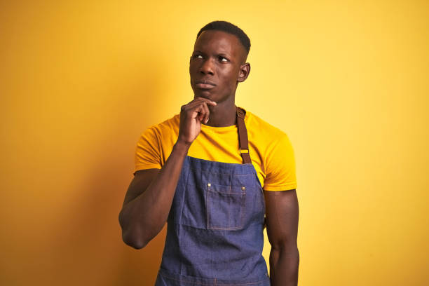 istock African american bartender man wearing apron standing over isolated yellow background with hand on chin thinking about question, pensive expression. Smiling with thoughtful face. Doubt concept. 1173780187