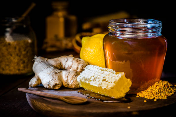 Honey jar with honeycomb and bee pollen, ginger and lime Front view of a honey jar surrounded by lemon, ginger, a honeycomb, a honey dipper and bee pollen. Objects are on a rustic cutting board and against black background. Low key DSLR photo taken with Canon EOS 6D Mark II and Canon EF 24-105 mm f/4L Honey stock pictures, royalty-free photos & images