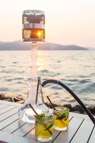 Two glasses of mojito with lime and mint with a hookah on a white table of an outdoor cafe by the sea at sunset. Travel and Vacation Concept