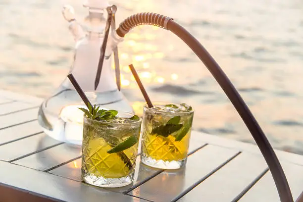 Two glasses of mojito with lime and mint with a hookah on a white table of an outdoor cafe by the sea at sunset. Travel and Vacation Concept