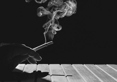 Sunlight and shadow on surface of man's hand holding cigarette with smoke on wooden table in dark background, black and white style