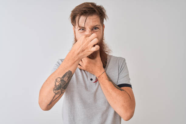 istock Young redhead irish man wearing grey polo standing over isolated white background smelling something stinky and disgusting, intolerable smell, holding breath with fingers on nose. Bad smells concept. 1173809921