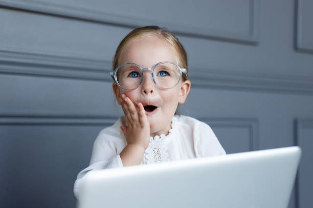 Portrait of cute little girl in eyeglasses behind laptop computer. Girl enjoying modern generation technologies, making mimic with her hands and mouth, isolated on a grey background. Copy space. stock photo