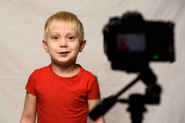 smiling blond boy in front of the camera lens. little video blogger. home studio - camera lens home video camera broadcasting imagens e fotografias de stock