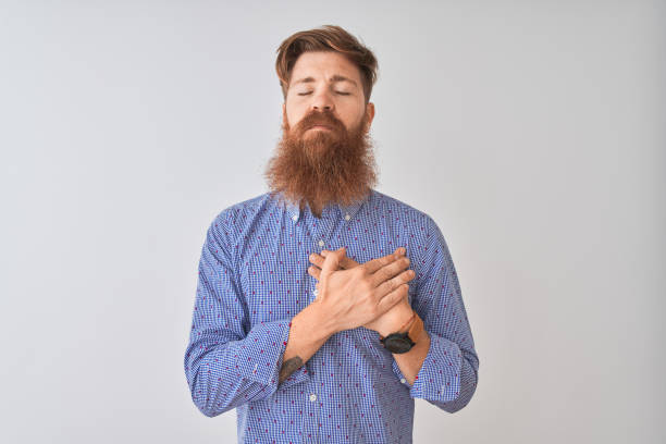 Young redhead irish man wearing casual shirt standing over isolated white background smiling with hands on chest with closed eyes and grateful gesture on face. Health concept. Young redhead irish man wearing casual shirt standing over isolated white background smiling with hands on chest with closed eyes and grateful gesture on face. Health concept. chest tattoos for men designs stock pictures, royalty-free photos & images