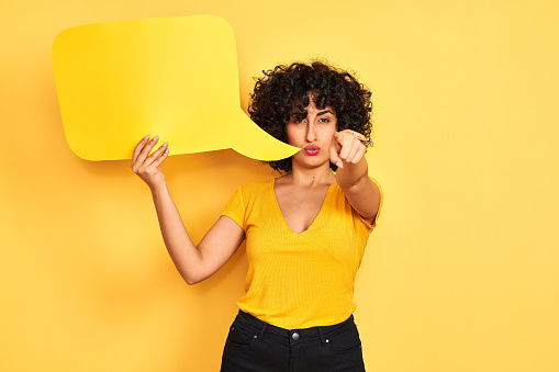 Young arab woman with curly hair holding speech bubble over isolated yellow background pointing with finger to the camera and to you, hand sign, positive and confident gesture from the front