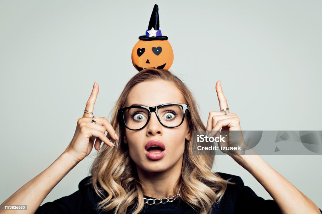 Surprised woman ready for halloween, stock photo Mid adult beautiful woman wearing black sweater and headband with pumpkin standing against grey background and staring at camera. Autumn Stock Photo