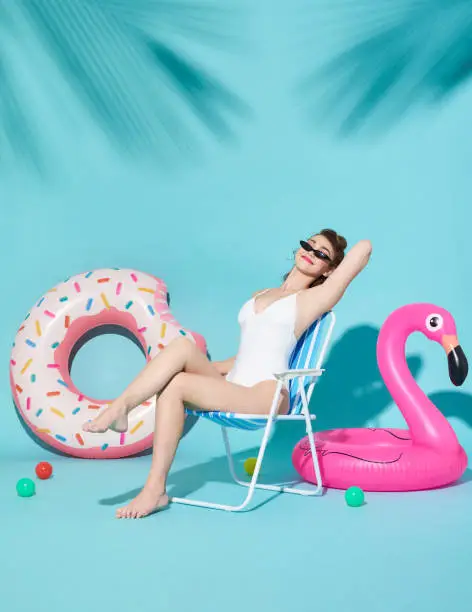 Cheerful attractive joyful delightful women dressed in nice swimwear sitting on a beach chair and rubber ring on bright blue background.