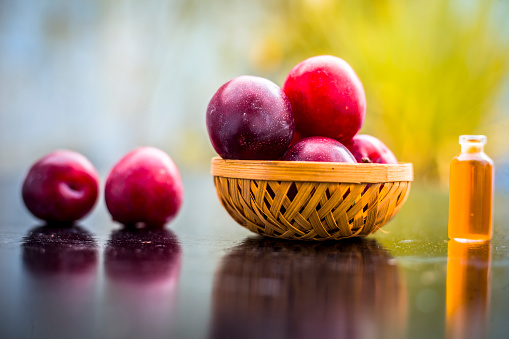Raw organic red ripe plums in a brown-colored basket on the wooden surface along with its extracted essential oil in a small transparent bottle with blurred background.