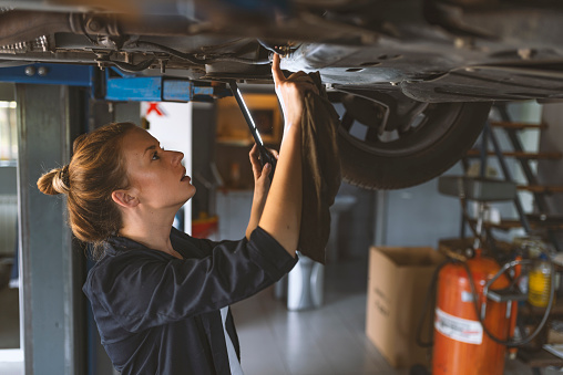 Female mechanic working on car tyre service. Portrait Of Female Auto Mechanic Working Underneath Car. Portrait of smiling young female mechanic inspecting a CV joing on a car in auto repair shop