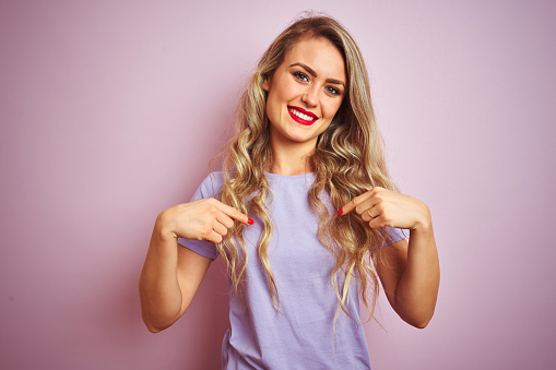 Young beautiful woman wearing purple t-shirt standing over pink isolated background looking confident with smile on face, pointing oneself with fingers proud and happy.
