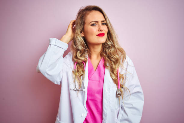 Young beautiful doctor woman using stethoscope over pink isolated background confuse and wonder about question. Uncertain with doubt, thinking with hand on head. Pensive concept.