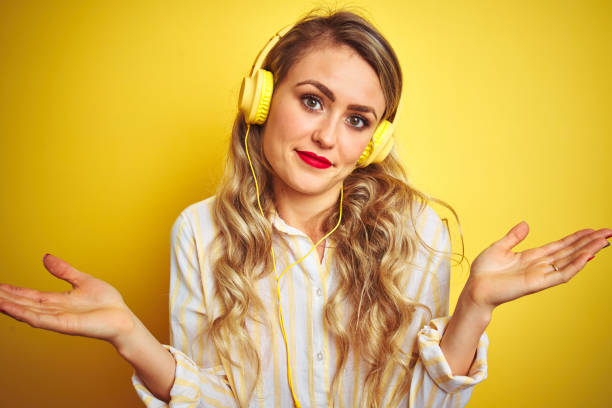 young beautiful woman listening to music using headphones over yellow isolated background clueless and confused expression with arms and hands raised. doubt concept. - head and shoulders audio imagens e fotografias de stock