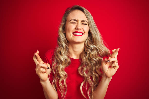 istock Young beautiful woman wearing basic t-shirt standing over red isolated background gesturing finger crossed smiling with hope and eyes closed. Luck and superstitious concept. 1173838480