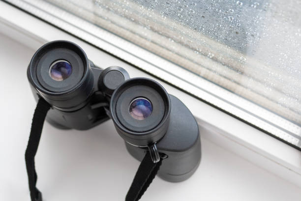 istock Closeup top view on a black binoculars for observing neighbors or nature, that stands on a white windowsill by the window with raindrops on the glass. Observation of the environment from the window 1173841087