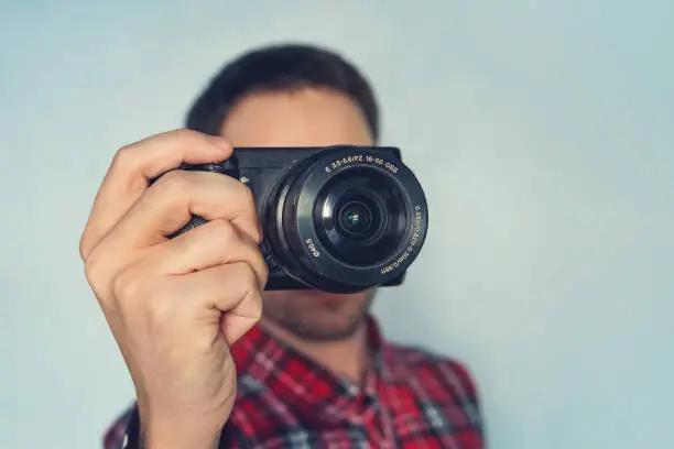 Close up photo of man in hat on blue background taking a photo with digital mirrorless camera. Young cute male photo reporter with a small camera in her hands. Paparazzi taking a picture.