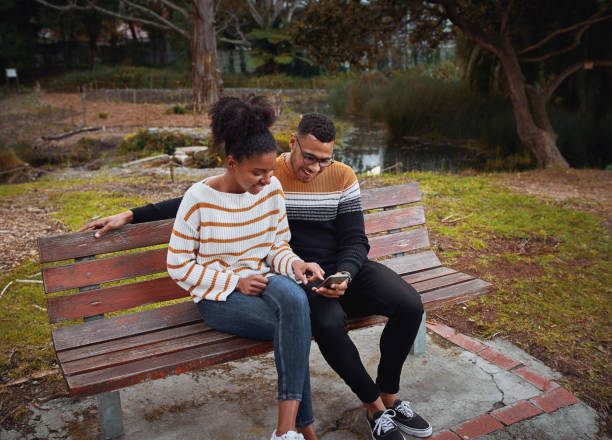 happy young african couple sitting together on bench near pond in park using mobile phone - using phone garden bench imagens e fotografias de stock