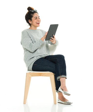 Full length / side view / profile view of 30-39 years old adult beautiful puerto rican ethnicity / latin american and hispanic ethnicity female / young women / one young woman only sitting / resting in front of white background wearing sweater / jeans / sunglasses who is smiling / happy / cheerful / cool attitude and using digital tablet / sitting on chair