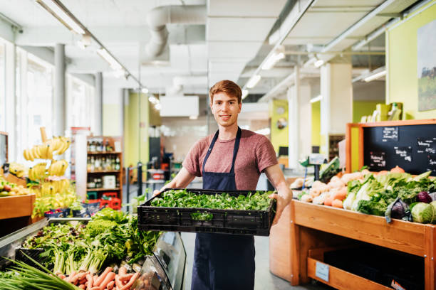 Portrait Of Young Supermarket Clerk Holding Crate Of Vegetables A portrait of a young supermarket clerk at work holding a crate of vegetables while stocking up the aisles. produce section stock pictures, royalty-free photos & images