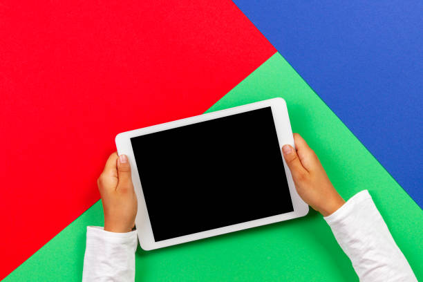 istock Kid hands holding white tablet computer on light green, blue and red background 1173897753
