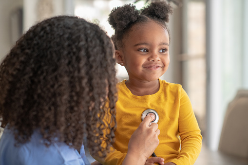 A little girl of African descent is at a medical appointment. She is sitting on an exam table. The female doctor is using a stethoscope to check the little girl's heart rate. The happy child is smiling.