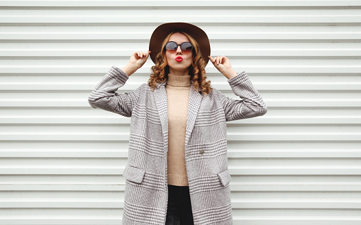 Stylish woman in checkered coat, round hat posing on city street over white wall background