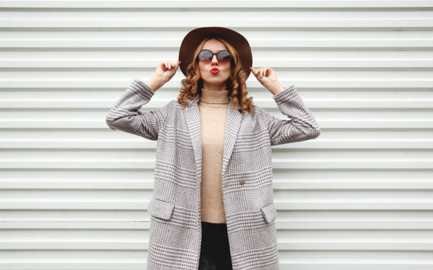 Stylish woman in checkered coat, round hat posing on city street over white wall background stock photo