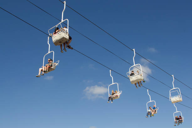 The selective focus of the skier's legs in the ski lift, the sun illuminates the six-seat ski lift. Below the cable car there is a ski slope and skis and a pine forest around.