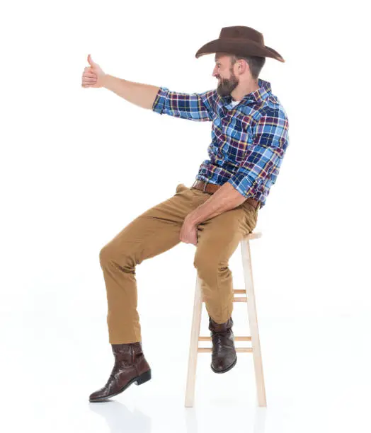 Photo of One man only / full length / side view / profile view of 30-39 years old adult handsome people caucasian young men / male cowboy sitting / resting in front of white background wearing cowboy boot / cowboy hat / hat who is smiling / happy / cheerful