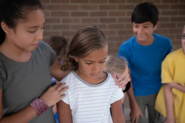 A mixed-race girl of 8 or 9 years looks dismayed as a group of children stand behind her and laugh as they bully her.
