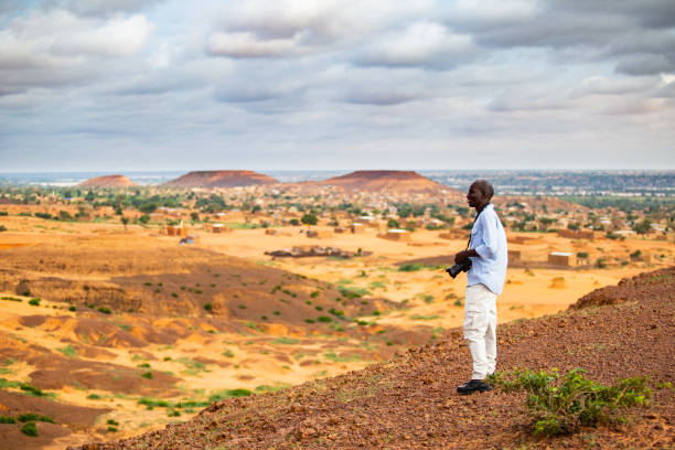 African man taking photos in the Sahel from a higher plateau having a traditional sahelian village in the background at the foothill of flat-top hills outside Niamey capital of Niger stock photo