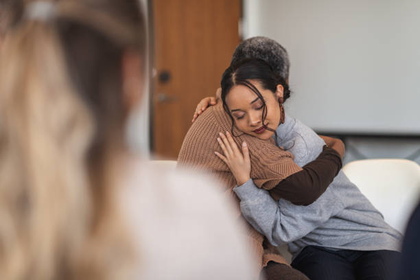 Supportive women hug while attending a group therapy session A young mixed-race woman hugs a mature adult black woman. They are sitting next to each other in a medical clinic. The two women are attending a group therapy session. They are showing support and kindness. woman comforting depressed friend stock pictures, royalty-free photos & images