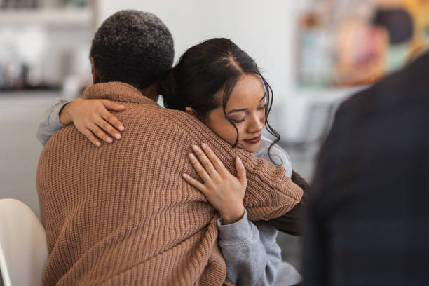 Supportive women hug while attending a group therapy session A young mixed-race woman hugs a mature adult black woman. They are sitting next to each other in a medical clinic. The two women are attending a group therapy session. They are showing support and kindness. group therapy photos stock pictures, royalty-free photos & images