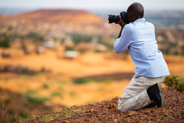 African man taking photos in the Sahel from a higher plateau having a traditional sahelian village in the background at the foothill of flat-top hills outside Niamey capital of Niger stock photo