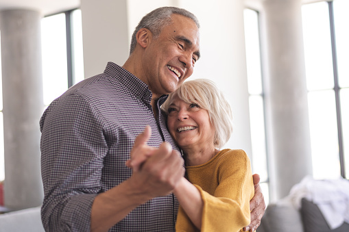A heterosexual senior couple is bonding. The husband is black and the wife is caucasian. They are dancing together indoors. The couple is smiling and enjoying this romantic moment together.