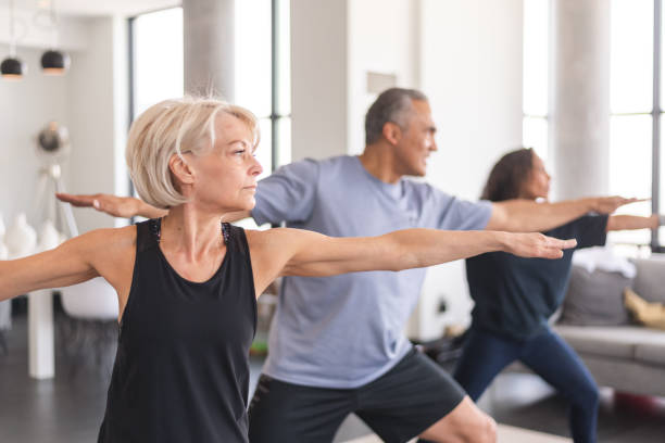 Active seniors in group fitness class A multi-ethnic group of seniors is attending a fitness class. They are indoors. The group is doing yoga. The individual in focus is a a beautiful and fit caucasian woman. community health center stock pictures, royalty-free photos & images