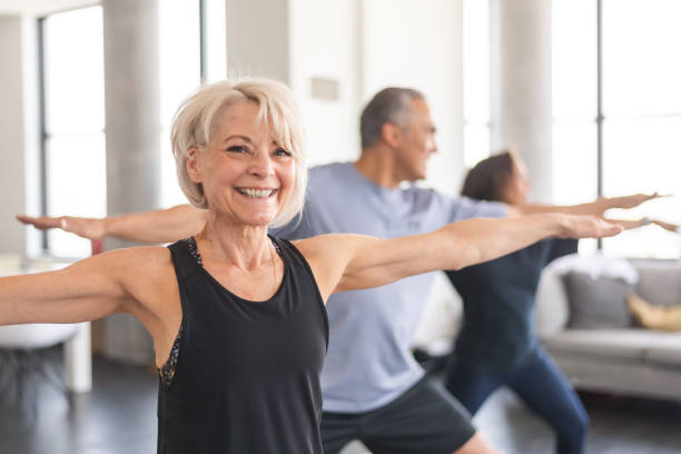 Active seniors in group fitness class A multi-ethnic group of seniors is attending a fitness class. They are indoors. The group is doing yoga. The individual in focus is a a beautiful and fit caucasian woman. She is smiling directly at the camera. young at heart stock pictures, royalty-free photos & images