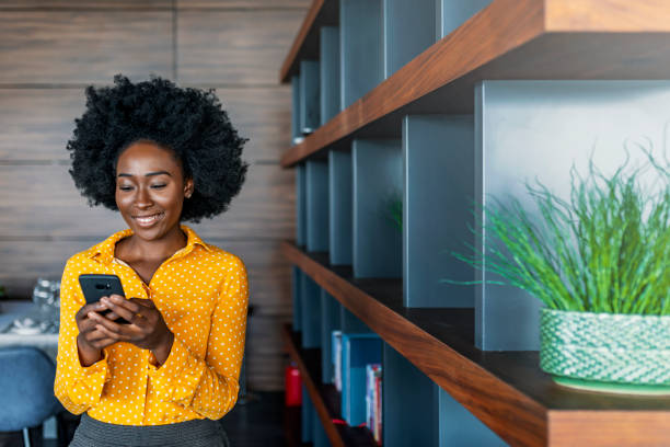 Business woman reading text message on mobile phone Quick message to friend. Attractive young African woman holding smart phone and looking at it while standing indoors. mobile phone text messaging telephone women stock pictures, royalty-free photos & images