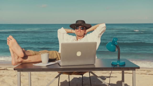 Young urban digital nomad of millennial generation z age sits on beach at resort or travel vacation destination, after work at laptop, resting putting his legs on table, remote office job, freelance