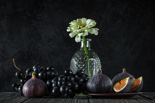 ripe whole and sliced figs with a bunch of black grapes and one flower in an empty decanter on old wooden boards against the background of a black textured concrete wall. elegant classic still life in the style of a dark photo. side view
