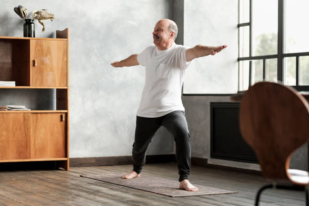 man standing in warrior two yoga pose practicing in living room Senior hispanic man standing in warrior two yoga pose practicing in living room alone warrior position stock pictures, royalty-free photos & images