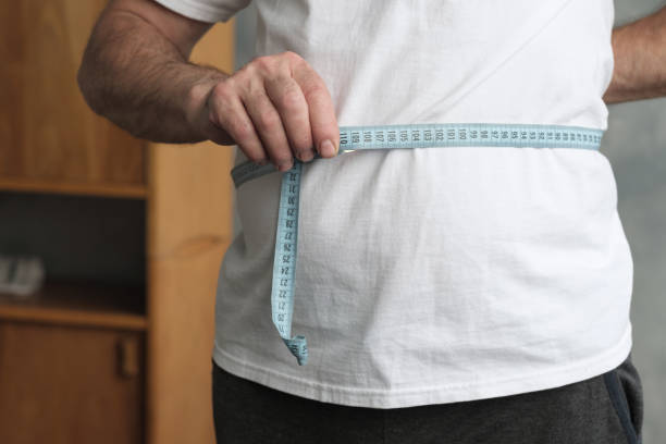 man measuring his belly with measurement tape standing in the living room. - measuring waist imagens e fotografias de stock