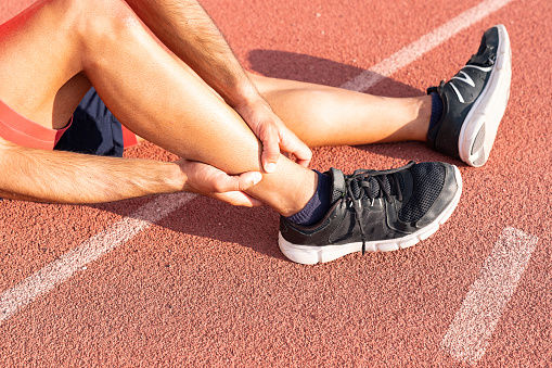 young sport man with strong athletic legs holding ankle with his hands in pain after suffering muscle injury during a running workout on Running Track. Healthcare and sport concept.