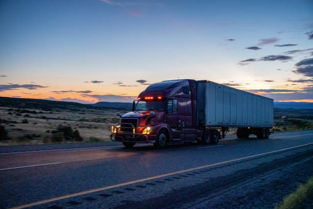 semi-truck speeding down a four lane highway with a dramatic sunset in the background and lights on - four lane highway stock-fotos und bilder
