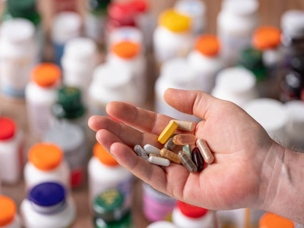 many nutritional health supplements and vitamins in capsules and tablets in a hand before consuming them, with many colorful food supplement and vitamin bottles out of focus in the background. - vibrant color healthcare and medicine healthy lifestyle vitamin pill imagens e fotografias de stock